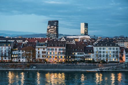 An aerial view of the historic riverfront buildings and the towering modern skyscrapers behind glow in the nights sky in the Wettstein district of Basel, Switzerland.