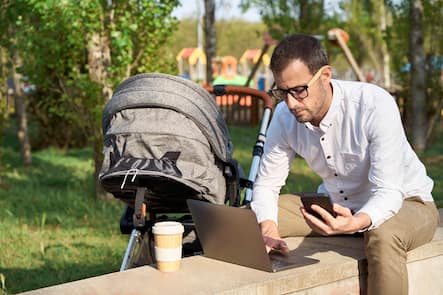 Male entrepreneur holding mobile phone while using laptop by baby stroller in park model released Symbolfoto VEGF04442