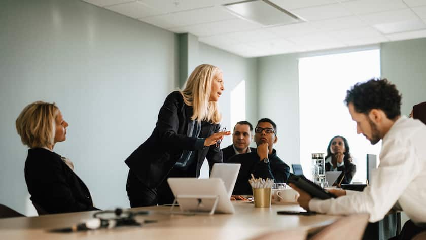 Eine Gruppe von Geschäftsleuten sitzt am Tisch. Eine mittelalte Frau steht, lehnt sich über den Tisch und erklärt etwas.