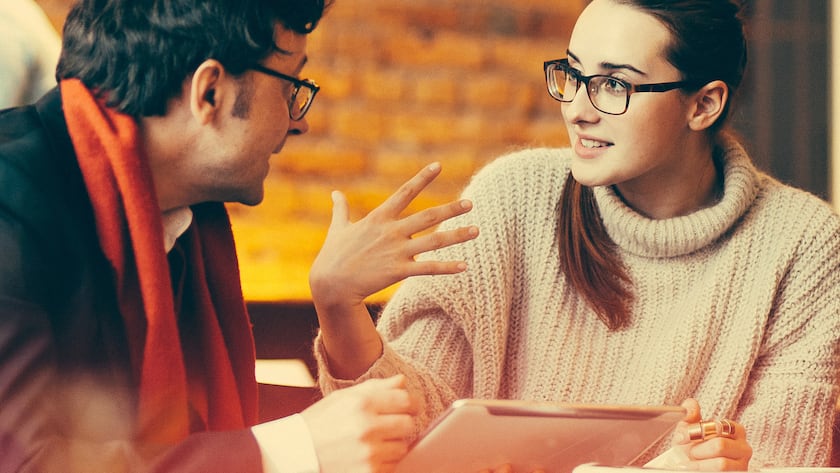 Eine junge Frau in Strickpullover hört einem Mann zu, der ihr in einer angesagten Location etwas erklärt.