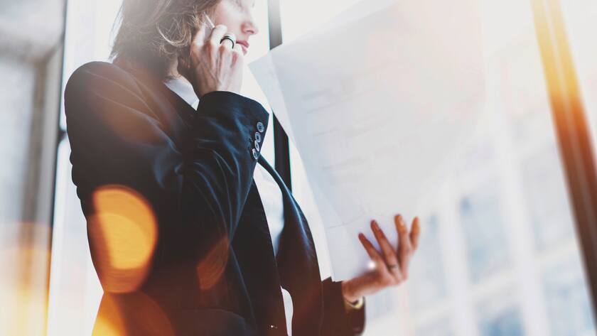 Photo business woman wearing white shirt, talking smartphone and holding documents in hands. Open space loft office. Panoramic windows background. Wide mockup