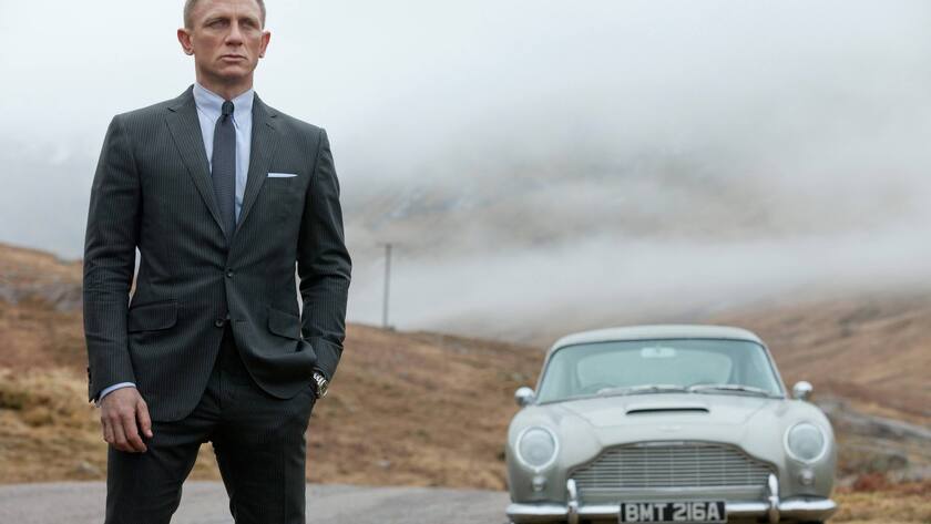 Daniel Craig steht im Vordergrund. Hinter ihm steht ein Aston Martin Db5.
