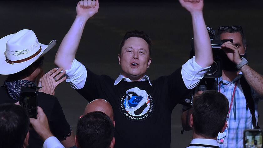 Elon Musk in einem SpaceX-Shirt reckt die Arme nach oben vor ihm stehen Journalistinnen und Kameramänner.