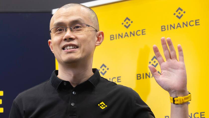 Changpeng Zhao aka CZ, der Gründer von Binance lächelt vor Wand mit Binance-Logo und winkt mir linker Hand.