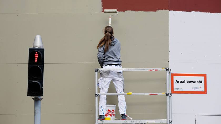 Eine Malerin auf einem Gerüst in Zürich malt ein Gebäude rot an, im Vordergrund ist eine Ampel zu sehen.