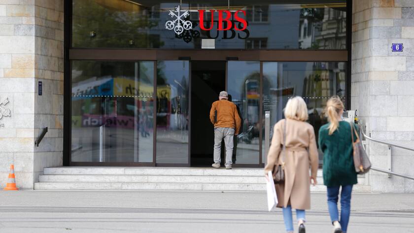 Zwei Frauen mit Rücken zur Kamera laufen auf UBS-Filiale zu mit Glastür. An der Glastür steht ein Mann in orangem Pullover.