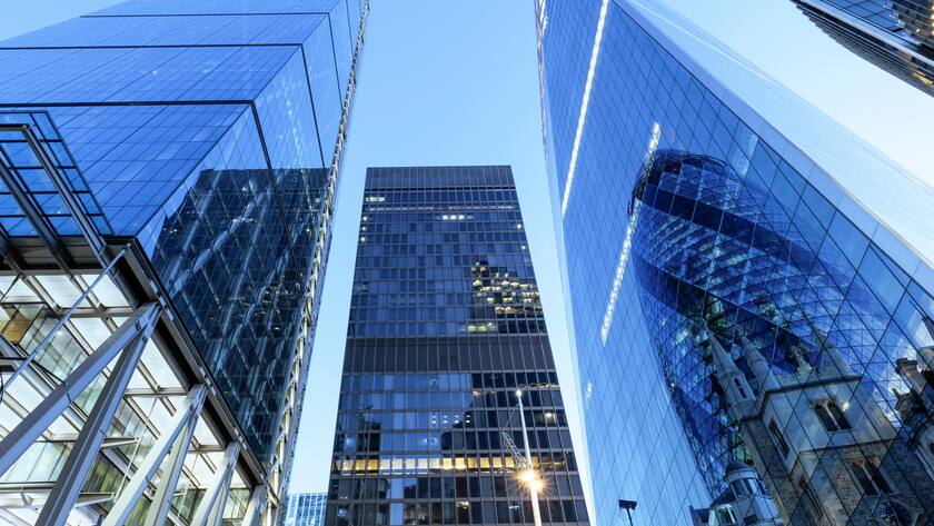 Attraktive Immobilienaktien UK, London, digital composite of modern office buildings in the city including the Cheesegrater, Aviva tower, Scalpel and the Gherkin, illuminated at twilight