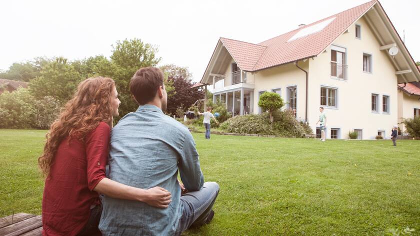 Frau und Mann sitzend im Garten ihres Einfamilienhauses, Kinder spielen im Garten