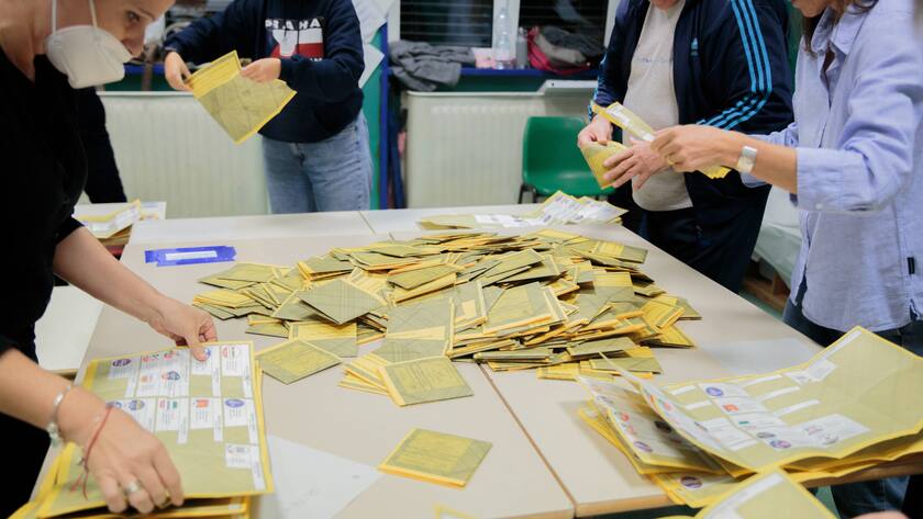 Mehrere Personen in einem Wahllokal stehen um einen Tisch mit Wahlcouverts.
