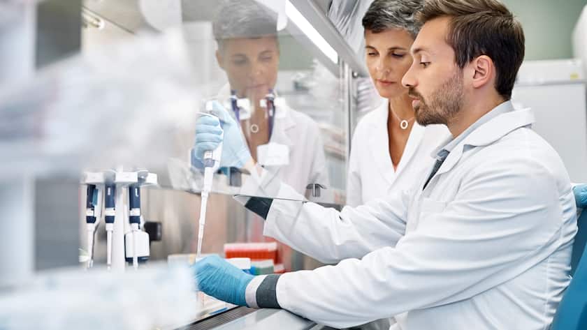 Female scientist looking at male colleague conducting cancer research. Confident doctors are working at biosafety cabinet. They are in laboratory.