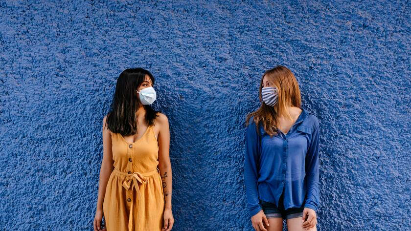 Zwei Frauen stehen vor einer blauen Wand, sehen sich an und tragen unterschiedliche Masken.