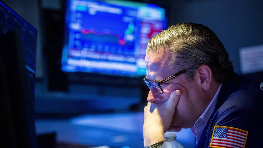 Mann mit Brille an US-Börse von Seite stützt Kinn in die Hand und blickt geradeaus in Bildschirm, der nicht zu sehen ist. Hinter ihm ist ein Bildschirm mit Kursen zu sehen.