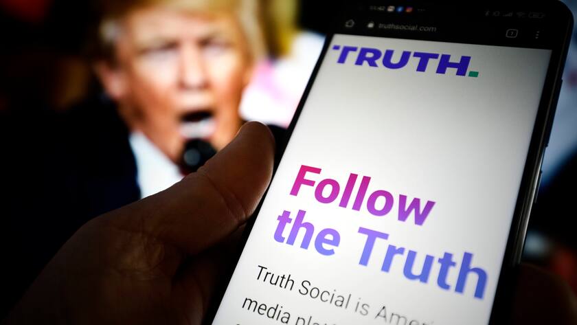 Das soziale Netzwerk Truth Social auf einem Handy-Bildschirm, im Hintergrund ein Foto des ehemaligen US-Präsidenten Donald Trump