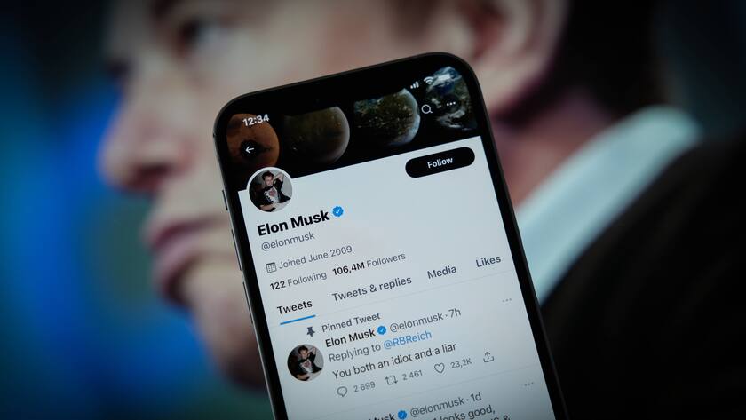 Elon Musk verschwommen im Hintergrund, davor ein Handy mit Elon Musks Twitter-Account drauf.