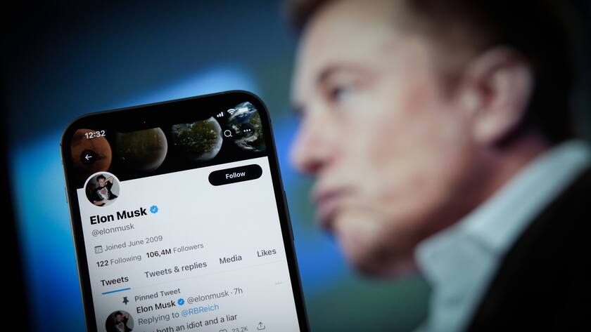 Elon Musk verschwommen im Hintergrund schaut nach links. Im Vordergrund ein Handy mit Musks Twitter-Profil drauf.