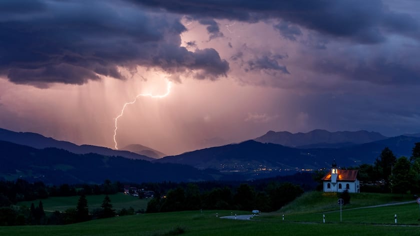Blitze entladen sich über den Alpen während eines aufziehenden Gewitters am Abend hinter der St. Hubertus Kapelle im bayerischen Scheidegg.
