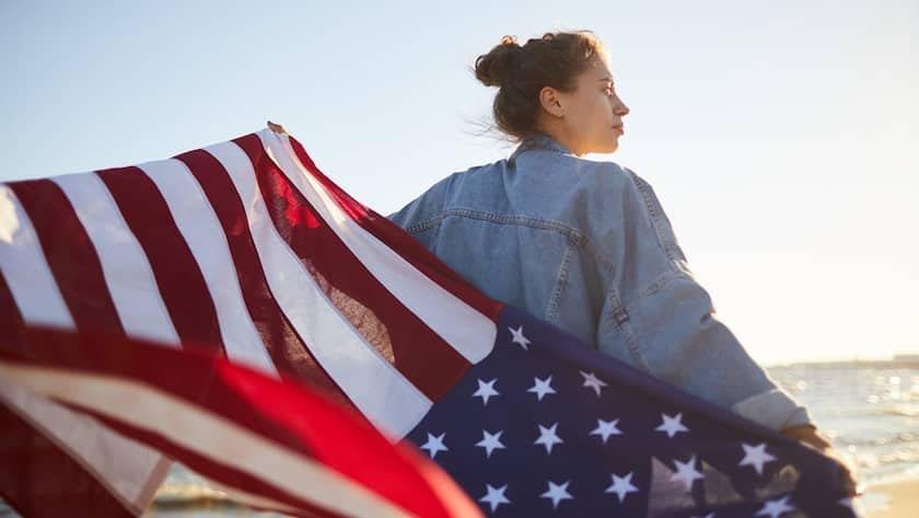 Rear view of introspective teenage girl in denim jacket outstretching arms with American flag and looking into distance while thinking of America