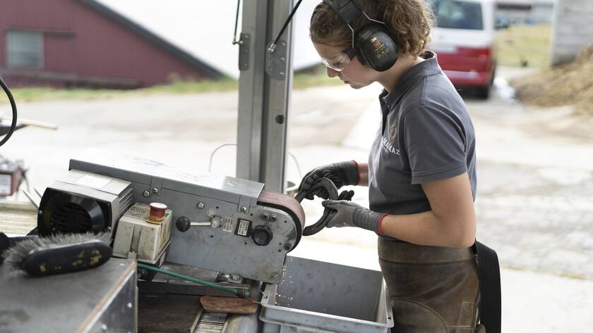 Eine Hufschmiedin in Ausbildung (EFZ) schleift ein Hufeisen an einer Schleifmaschine, aufgenommen am 16. September 2021 in Cronay Kanton Waadt. Die an der Bild-Dokumentation beteiligten HufschmiedInnen arbeiten im Hufschmied-Betrieb der Skalkaz Marechalerie Sarl. (KEYSTONE/Gaetan Bally)