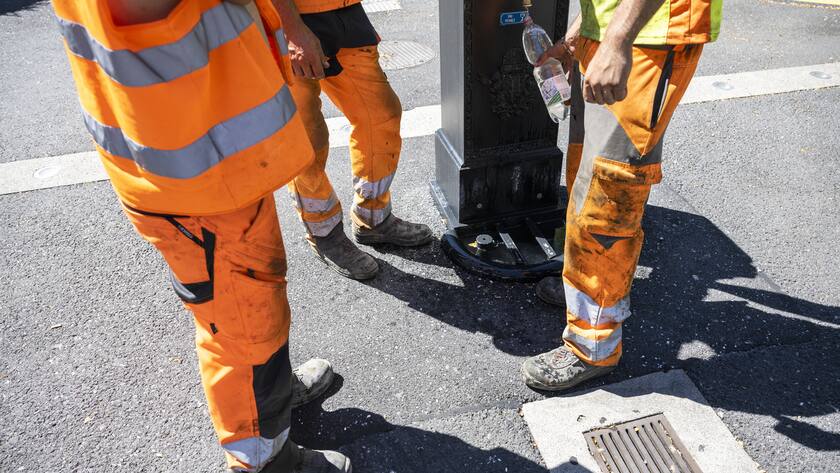 Drei Bauarbeiter stehen nebeneinander, es sind nur die Beine und Füsse zu sehen. Einer lässt Wasser in eine Petflasche laufen.