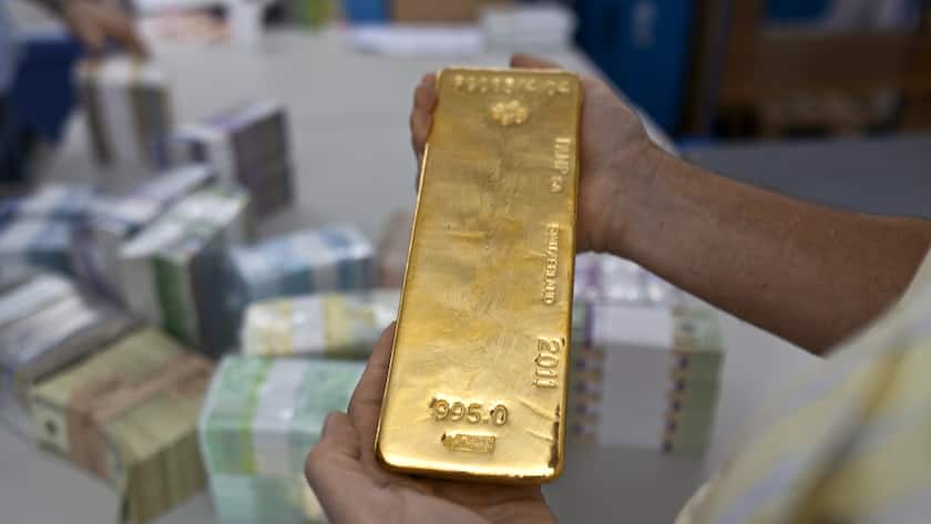 Eine Person hält einen Goldbarren in der Hand, im Hintergrund ist verschwommen Geld zu sehen.