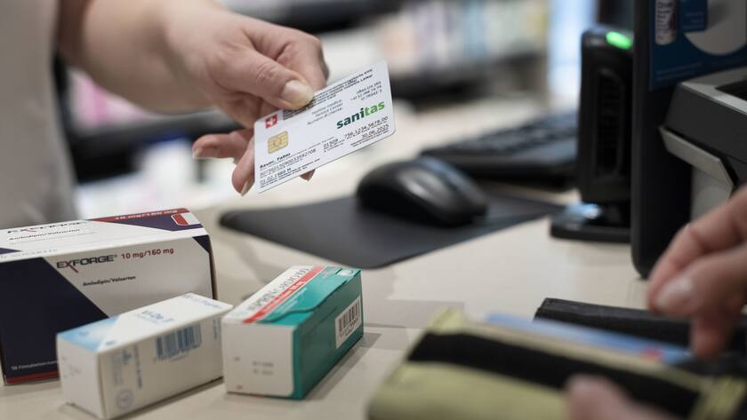In der Apotheke: Eine Hand hält eine Sanitas-Krankenkasse, gegenüber die Hand der Kundin am Portemonnaie. Dazwischen liegen Medikamente auf dem Tisch.