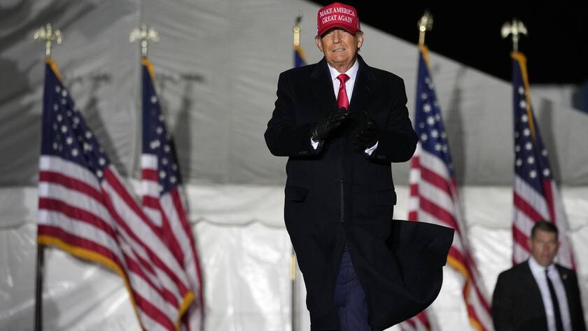 Donald Trump in schwarzem Mantel mit rotem "make america great again" Käppchen vor mehreren Flaggen.