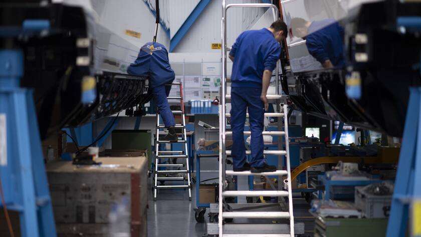 Blick in die Fertigung der Stadler Rail. Zwei Personen in blauen Arbeitskleidern arbeiten an Zügen.