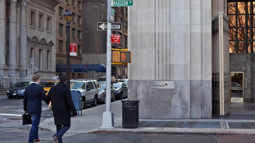 Zwei Fussgänger in New York von hinten laufen an Credit Suisse Gebäude vorbei neben einem Rotlicht.