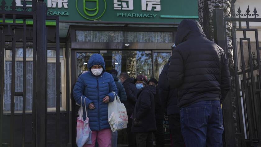 Eine Frau trägt eine Tasche mit Arzneien gegen Erkältung und Fieber, während andere Menschen vor der Apotheke Schlange stehen.