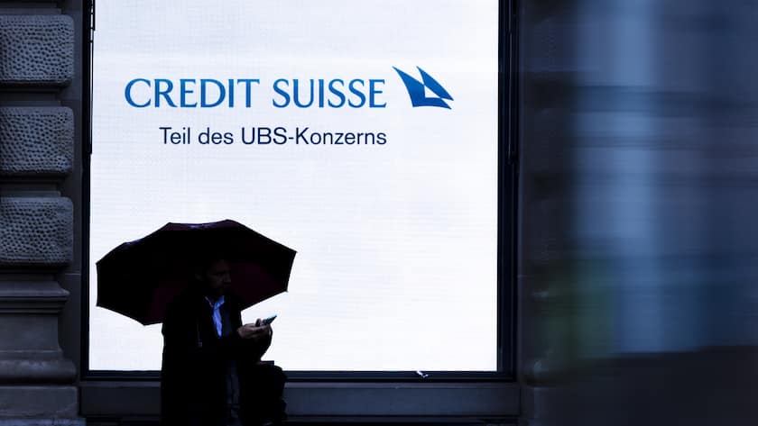 A person holding an umbrella is silhouetted against a screen of Swiss bank Credit Suisse with the words "Part of the UBS Group" at Zurich's Paradeplatz on Monday, June 3, 2024 in Zurich, Switzerland. (KEYSTONE/Michael Buholzer)