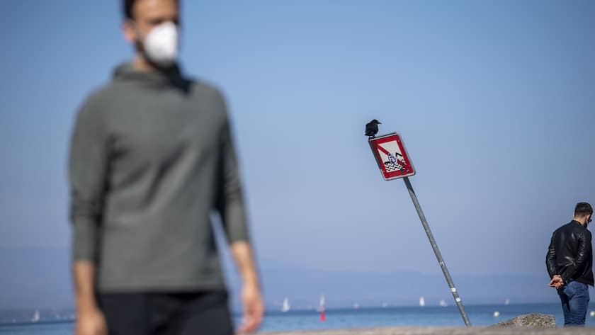 Une personne passe avec son masque de protection devant une panneau ou un corbeau s'est installe, les gens malgre les mesures d'eloignement social et le rassemblement de plus de 5 personnes, pendant l'etat d'urgence suisse en raison du coronavirus COVID-19, profitent de prendre l'air et le soleil, ce dimanche 5 avril 2020 a Geneve (KEYSTONE/Martial Trezzini)