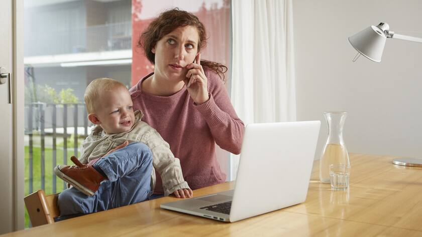 Eine Frau sitzt vor einem Laptop. Im rechten Arm hält sie ihr strampelndes Kleinkind, mit der linken Hand telefoniert sie am Handy.