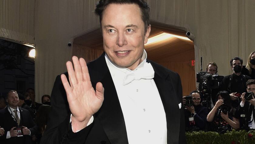Elon Musk im Tux winkt mit der rechten Hand und lächelt.