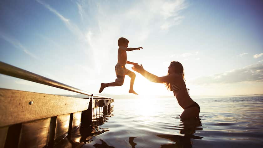 Ein junge springt von einem Steg in die Arme seiner Mutter, die im untiefen See steht und ihn auffängt.
