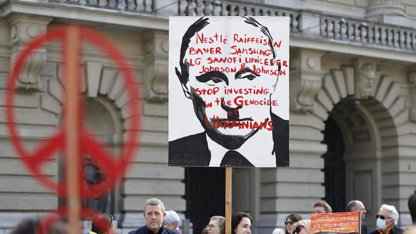 A protester holds a banners reading "Nestle Raiffeisen Bayer Samsung LG Sanofi Unilver Johnson & Johnson - Stop investing in the genocide of Ukrainians" during a demonstration against the Russian invasion of Ukraine in front of the Swiss parliament building in Bern, Switzerland, Saturday, March 19, 2022. (KEYSTONE/Peter Klaunzer)