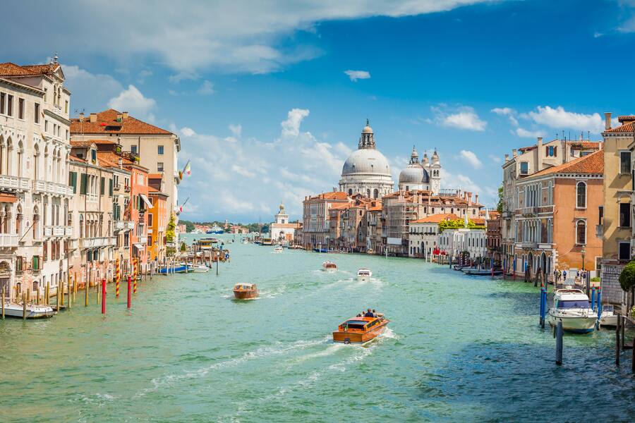 An iconic view from Accademia Bridge (Italian: Ponte dell'Accademia) in Venice. Numerous boats and vessels are seen cruising along the Grand Canal on a bright sunny day. Santa Maria Della Salute is seen in background.