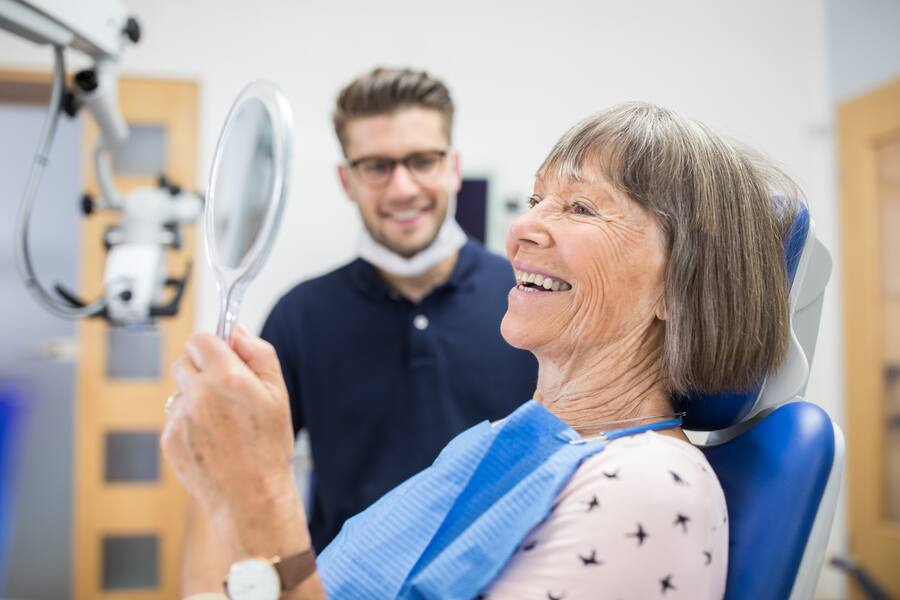 Smiling elderly patient checking her teeth into a hand mirror after her dental treatment. Senior patient checking her teeth after treatment at dental clinic.